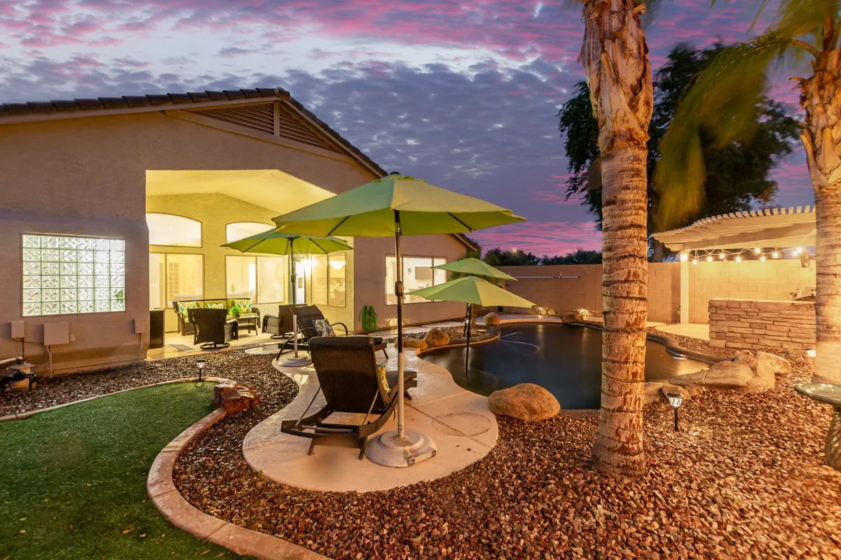 This exterior image features a sparkling swimming pool surrounded by green umbrellas, comfortable patio seating, and lush landscaping with stones and grass. The inviting setup is ideal for relaxing, entertaining guests, and enjoying sunset views, with outdoor lighting enhancing the tranquil atmosphere.