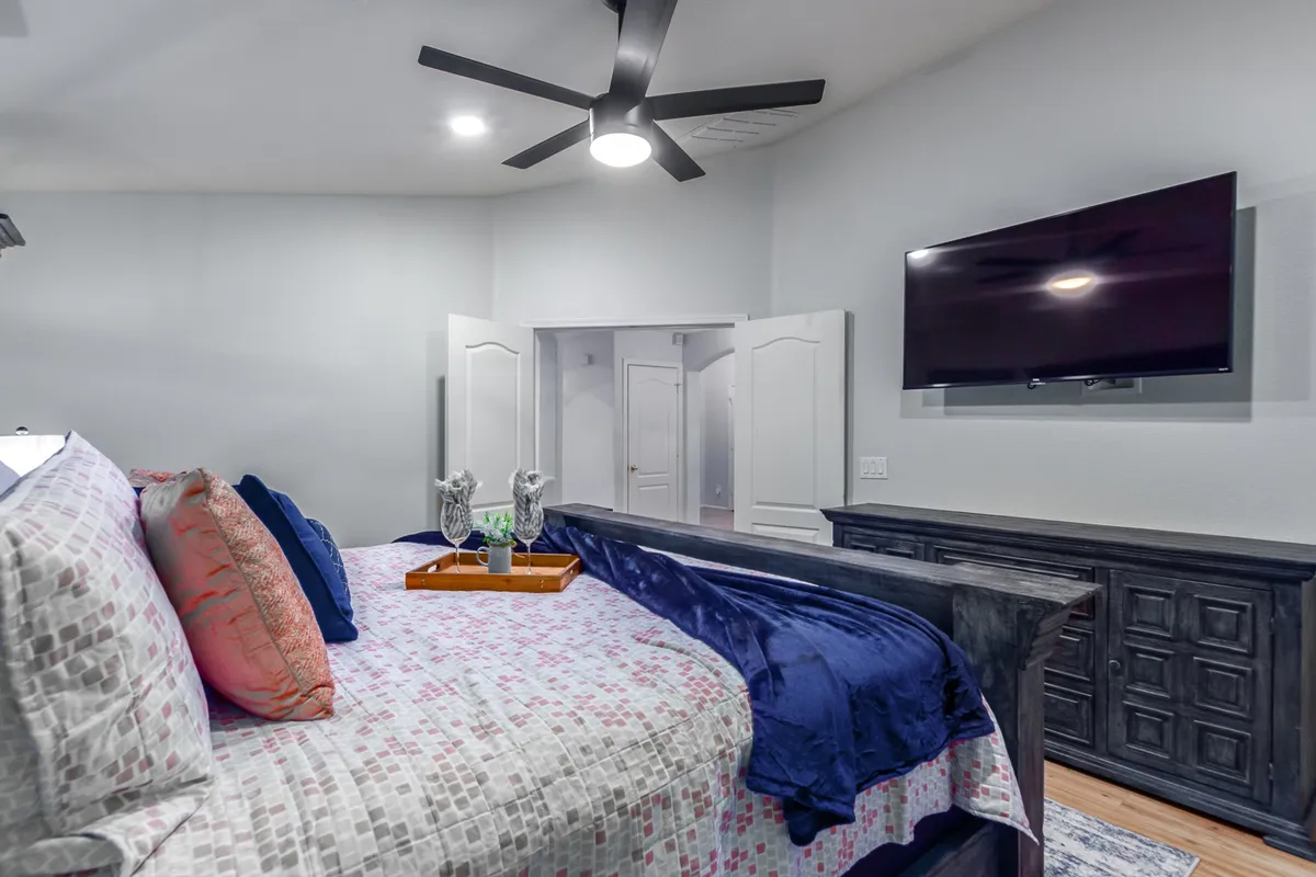 A serene bedroom with a bed dressed in decorative pillows, a wall-mounted television, ceiling fan, and a dresser adorned with decorative elements. The atmosphere is calm and inviting, perfect for relaxation or enjoying a quiet morning.