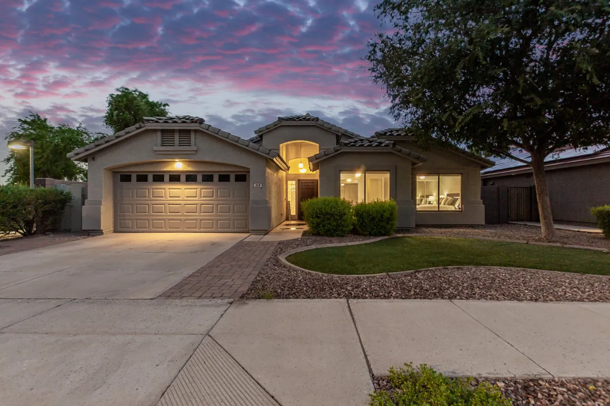 The image features the home's front entrance, garage, a landscaped yard, and windows glowing with warm light. The scene is set at dusk, suggesting both arrival and anticipation for evening activities.