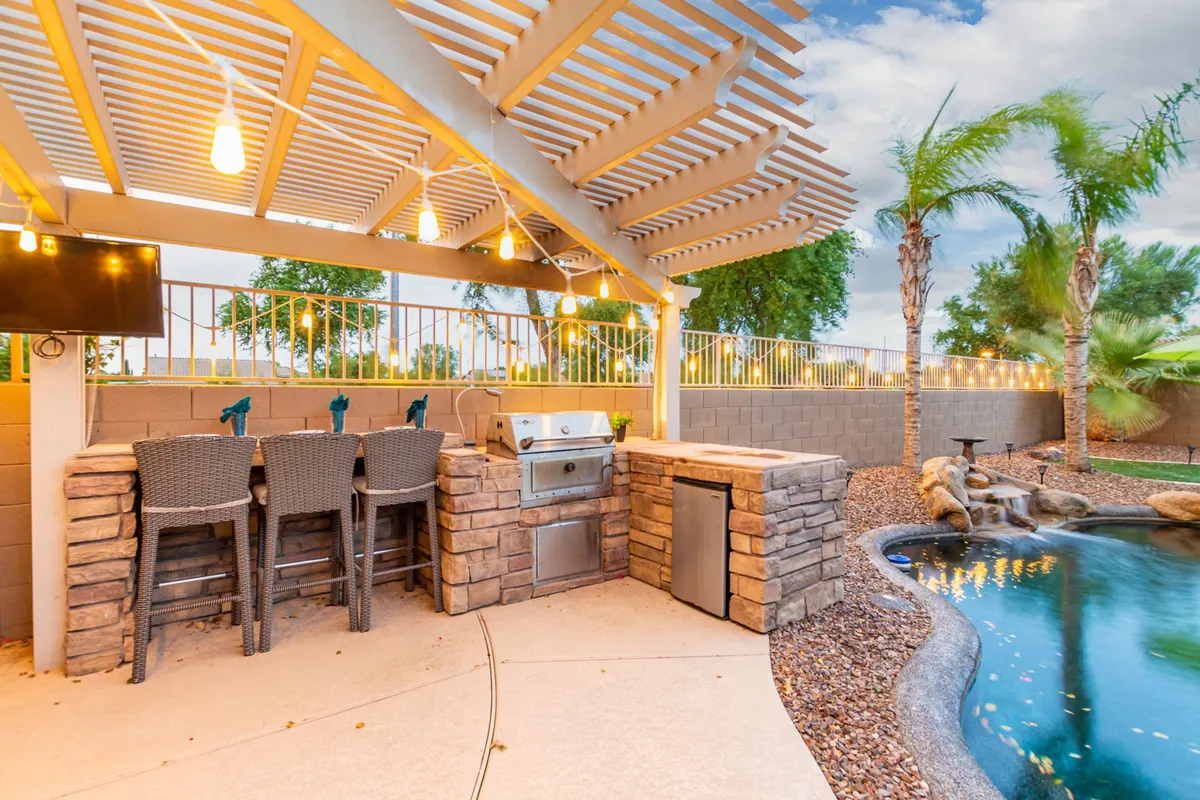 A festive exterior scene featuring a fully-equipped outdoor kitchen with a grill, bar seating, an adjacent swimming pool, and cozy seating under string lights. The area is designed for barbecuing, poolside lounging, and evening gatherings.