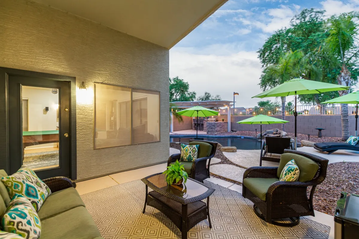 This exterior scene features a tranquil outdoor seating area adjacent to a swimming pool, complete with green umbrellas and decorative pillows. The setup invites social gatherings, outdoor meals, and relaxation, with the pool and patio signaling the connection between indoor and outdoor living.