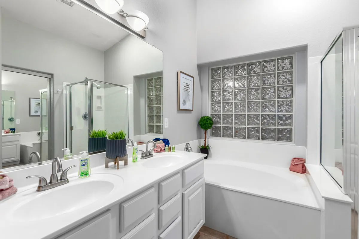 A spa-inspired bathroom with dual sinks, a relaxing bathtub, glass shower, framed artwork, and decorative greenery. Sunlight filters through glass blocks, creating a serene and refreshing ambiance.