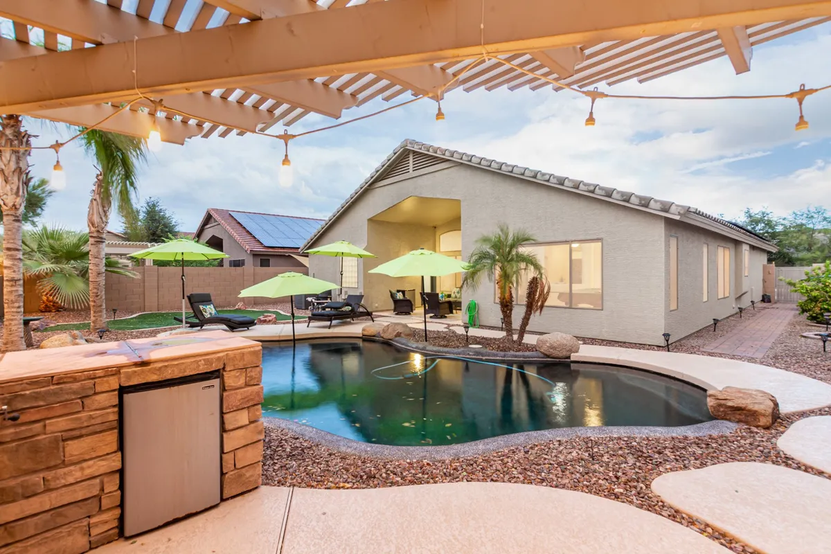 A lively backyard scene featuring a heated and chilled swimming pool surrounded by green umbrellas, an outdoor seating area, and a stylish stone bar. The area is set up for swimming, sunbathing, and socializing, with ample space for groups to gather and enjoy the outdoors.