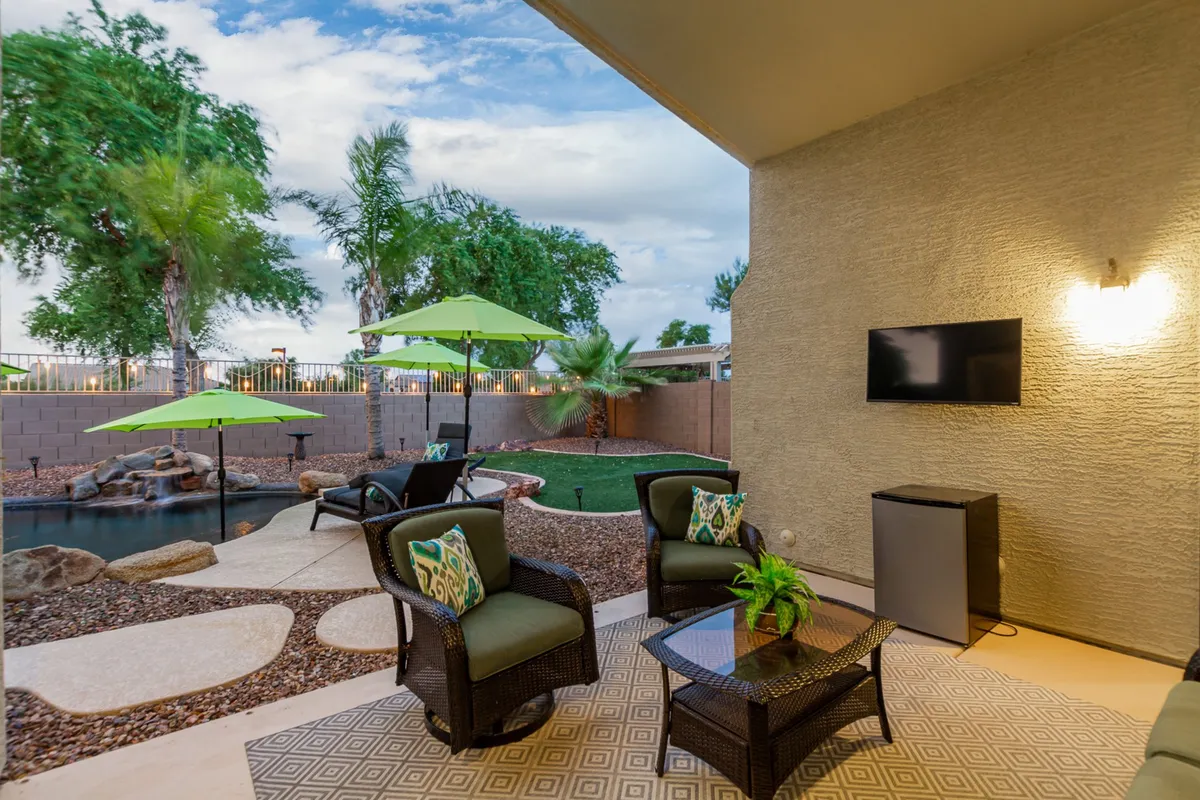 A serene patio area with comfortable outdoor seating, green umbrellas for shade, a sparkling swimming pool, and a TV mounted on the wall. The inviting space is perfect for relaxing, entertaining, or watching TV outdoors, with sunset views possible from the patio.