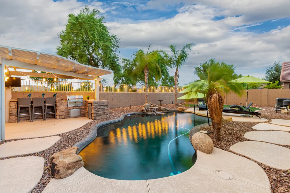 This image captures a sparkling swimming pool surrounded by palm trees, an outdoor bar area, landscaped stones, and ample outdoor seating. People are seen swimming, barbecuing, and relaxing under umbrellas, creating a lively, resort-like ambiance. String lights add a festive touch as the sun sets in the background, providing a warm, welcoming glow.