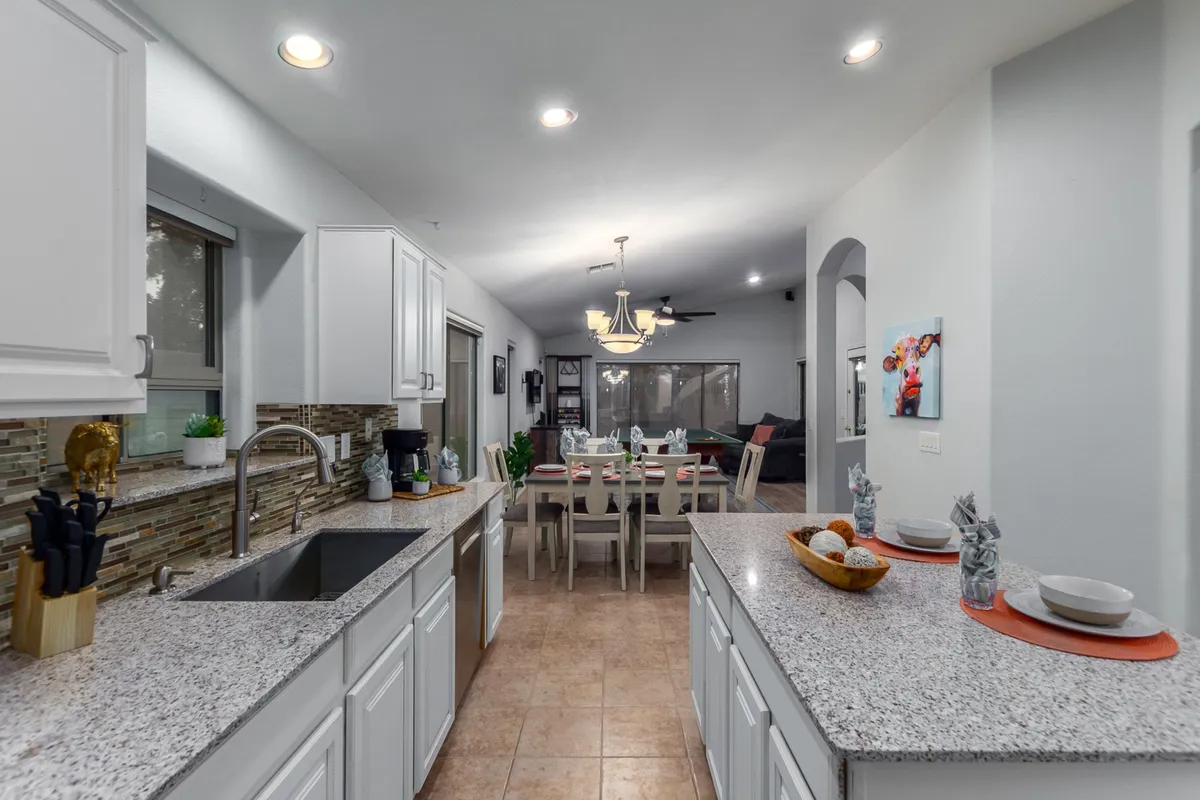 A spacious, open-concept kitchen and dining area filled with warm light. The image features a prominent kitchen island, an adjacent dining area set for a group, and stylish decorative touches throughout. Large windows allow ample natural light to fill the space, creating a welcoming environment for cooking and gathering.