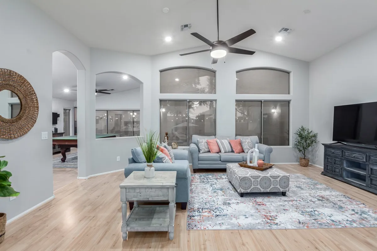 A bright, welcoming living room with a comfortable sofa adorned with decorative pillows, a central coffee table with accent items, a sleek TV unit, and expansive windows flooding the space with natural light. The arrangement invites both relaxation and social gatherings.