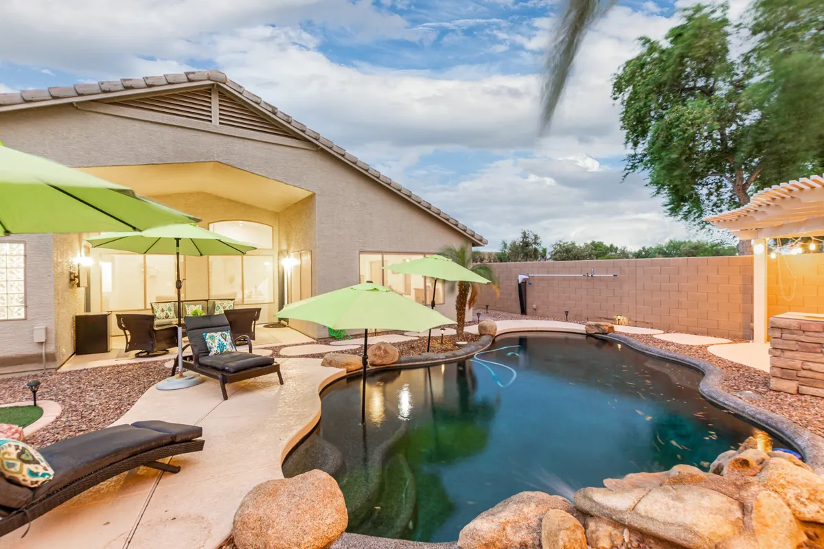 A tranquil poolside scene with lounge chairs, green umbrellas, rock formations, and vibrant landscaping. The setup is ideal for sunbathing, swimming, or gathering with family and friends in a peaceful backyard retreat.