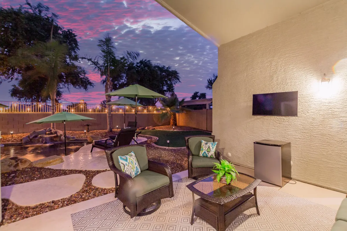 The photo showcases a cozy patio seating area with lush green umbrellas, a clear view of the pool, decorative plants, and an outdoor television. The scene is tranquil and social, ideal for watching the sunset or hosting an outdoor movie night. The inviting setup emphasizes relaxation and togetherness in the fresh air.