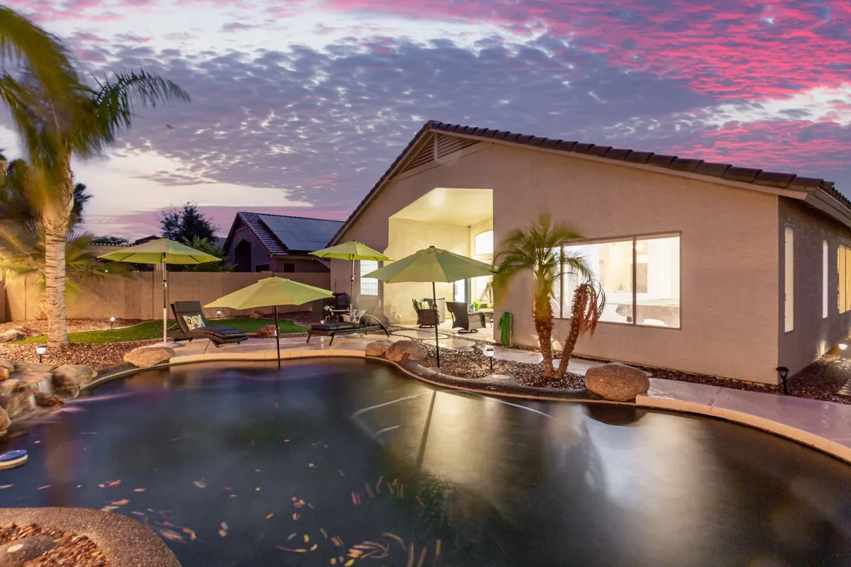 An exterior shot of the swimming pool area framed by palm trees and green umbrellas, with the house's exterior visible. The scene captures a calm, inviting atmosphere perfect for enjoying sunset views or social gatherings by the water.