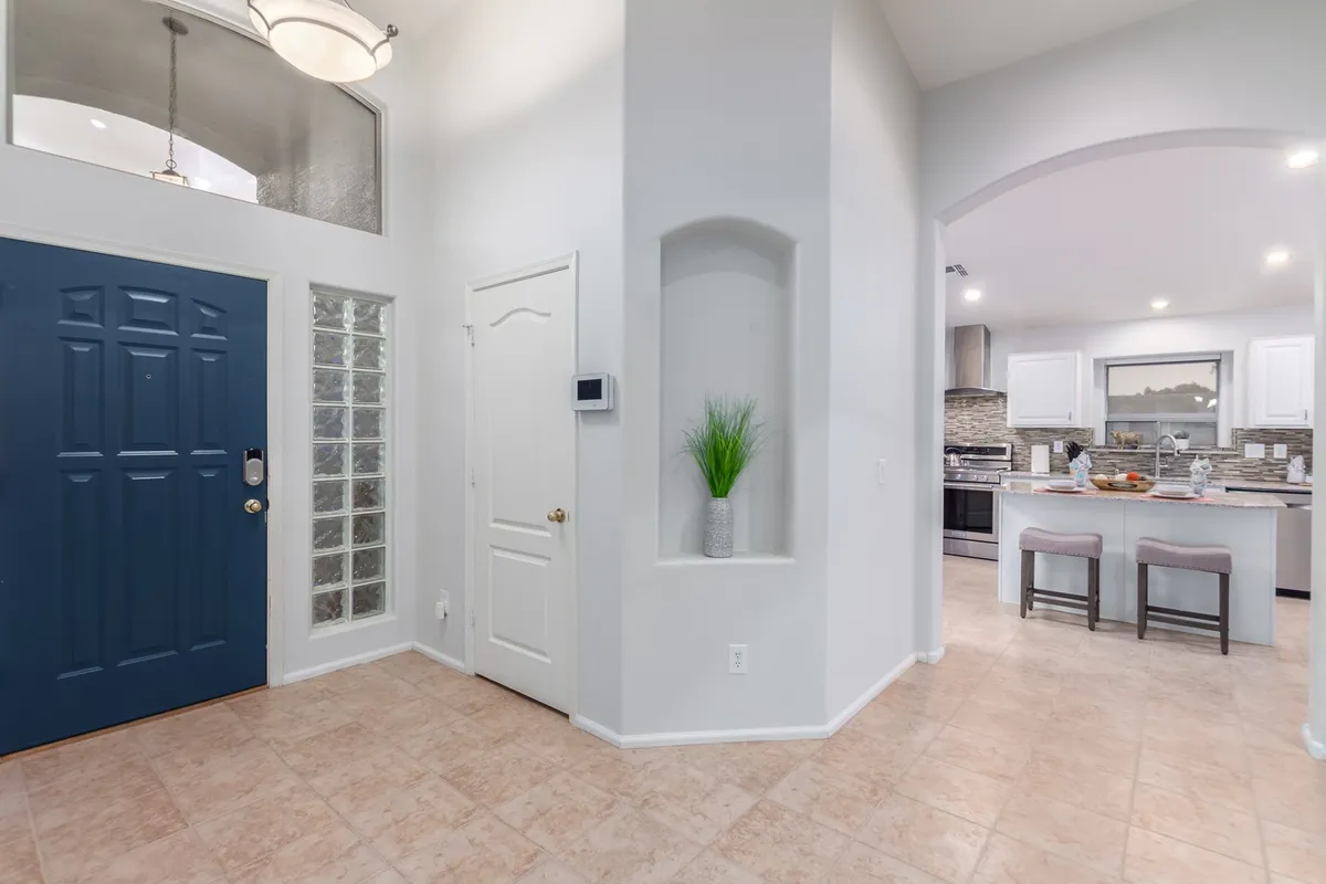 This image captures the blue front door opening to an arched entryway with a clear view into the kitchen and living space. A decorative plant adds warmth, and the open-plan layout is visible, illustrating how the indoor space flows smoothly for gatherings and activities.